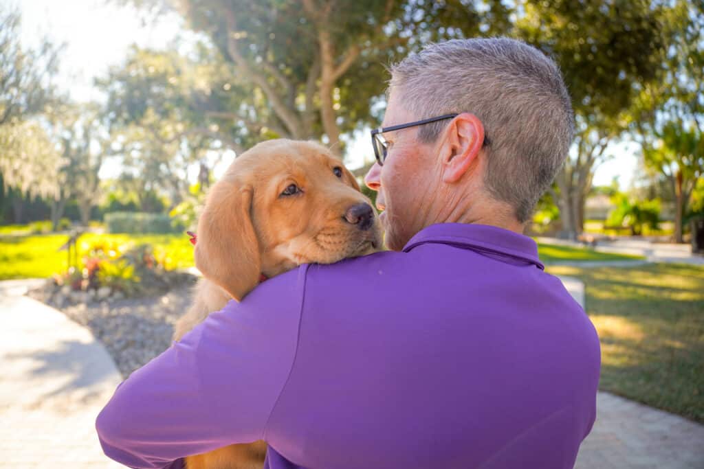 A pack member in a purple shirt holds a golden retriever puppy over their shoulder while standing outdoors in a park.