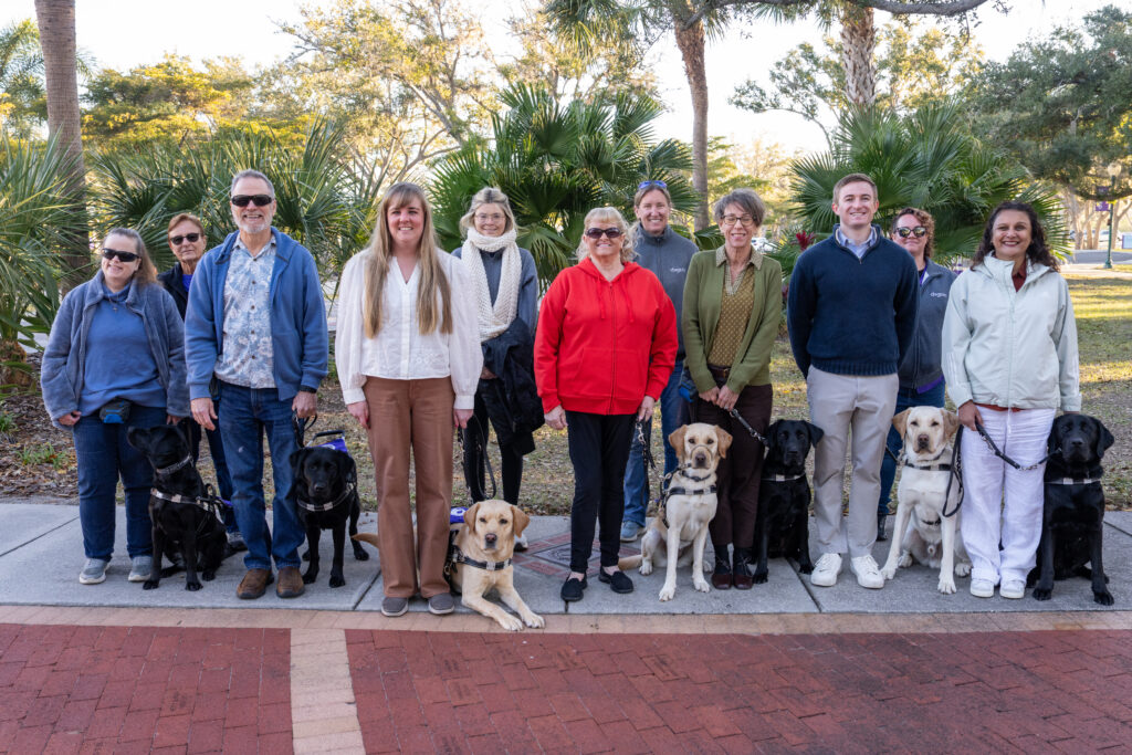 Group of eleven adults standing outdoors with seven Labrador Retrievers on leash, posing together on a sunny day on campus with trees in the background.
