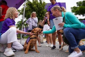 Two women kneel to pet a brown puppy wearing a purple trainee vest while others smile and gather around at an outdoor Dogs Inc event.