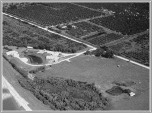 Historic black-and-white aerial photograph of the original Dogs Inc campus, showing buildings, ponds, open fields, and surrounding groves.