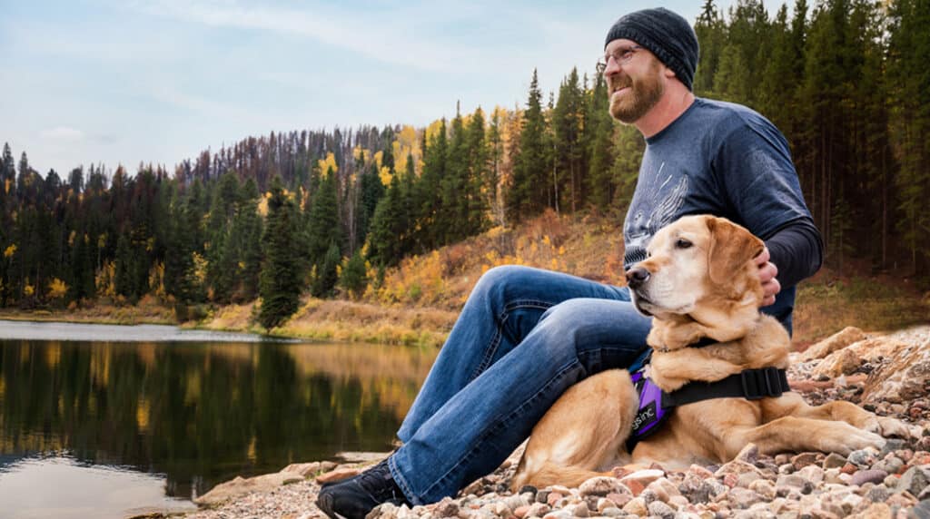 Man sitting on a rocky lakeshore with a yellow dog in a purple Dogs Inc vest, both looking out over a calm lake surrounded by forest trees.