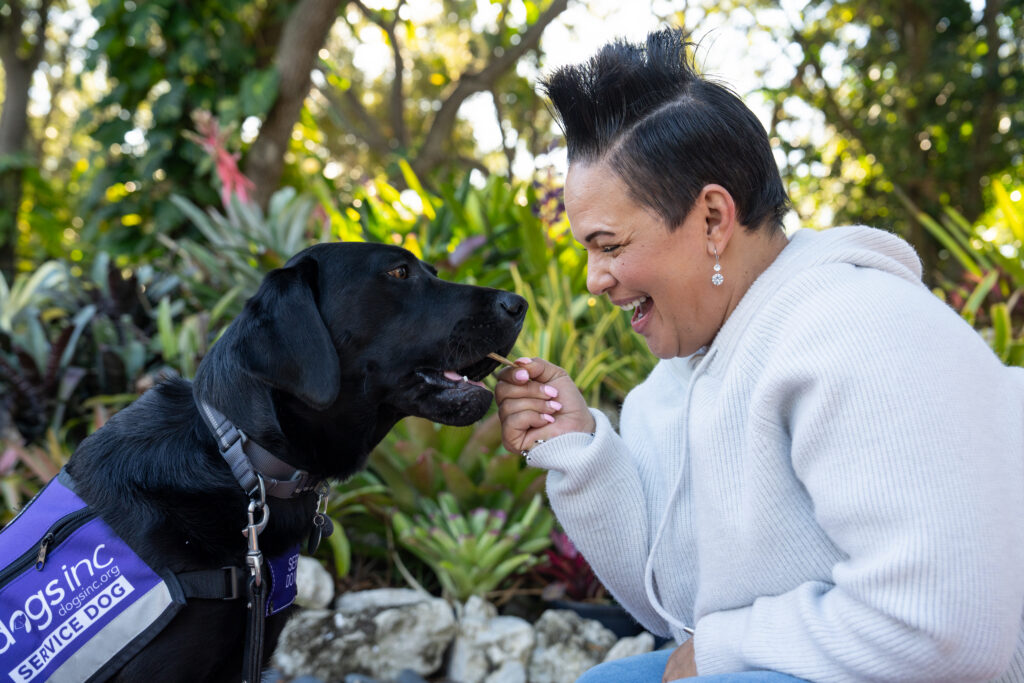 Woman smiles while giving a treat to a black Labrador Retriever wearing a purple Dogs Inc vest labeled "service dog" in an outdoor garden setting.