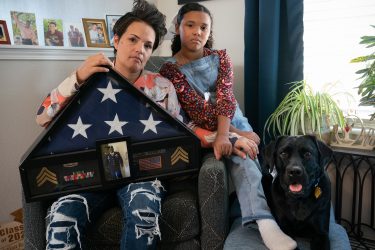 A mom and young girl sit and hold a folded American flag with a black lab Goldstar dog lays next to them