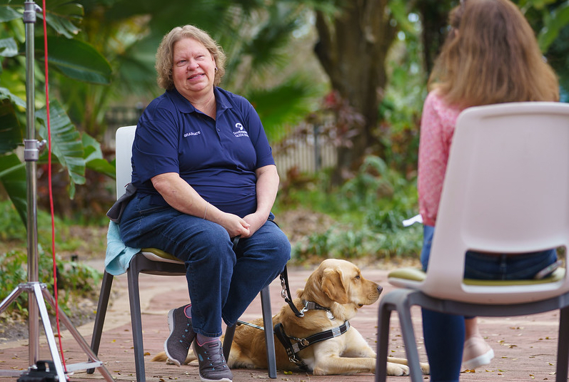 Woman sits in a chair with yellow lab guide dog in harness laying next to her with a woman sitting across from them