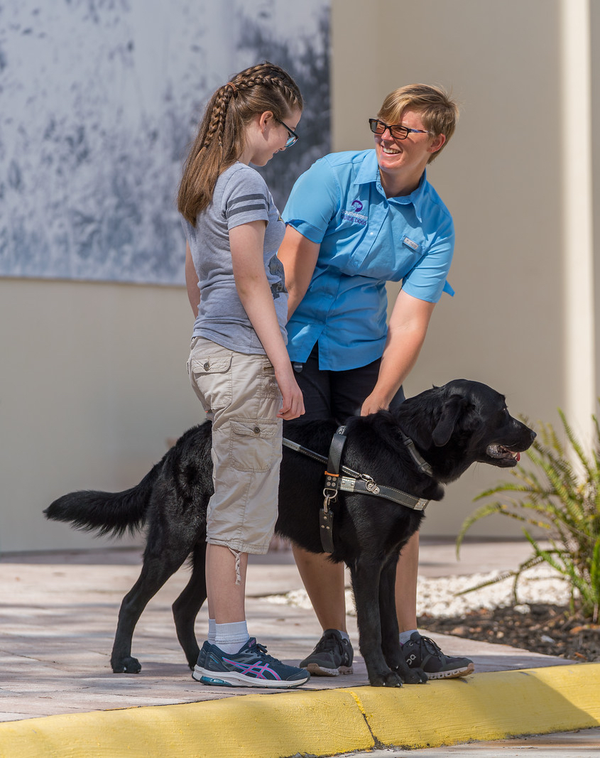 Teen girl stands at curb with black guide dog in harness while trainer talks
