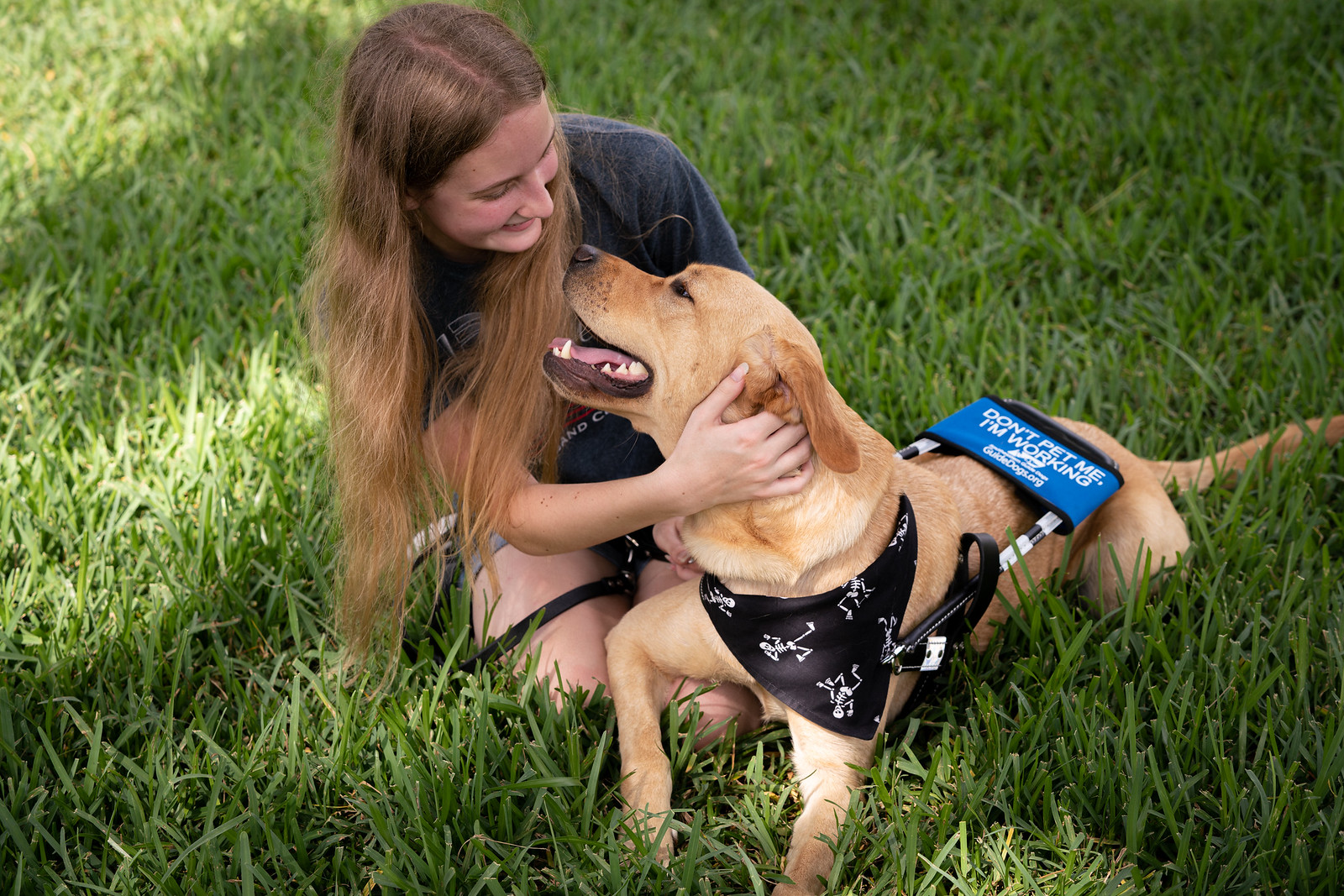 Teen girl sits in grass while her guide dog in harness lays in grass wearing a halloween skeleton bandana