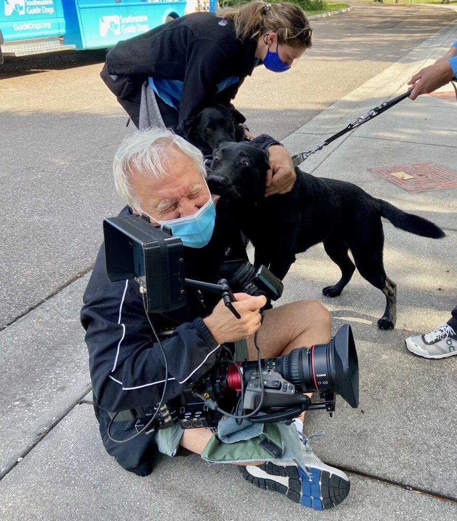 A camera man sits on the concrete smiling as a black lab kissing his ear