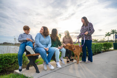 A family of five sits on an outdoor bench with their yellow Labrador on a purple leash.