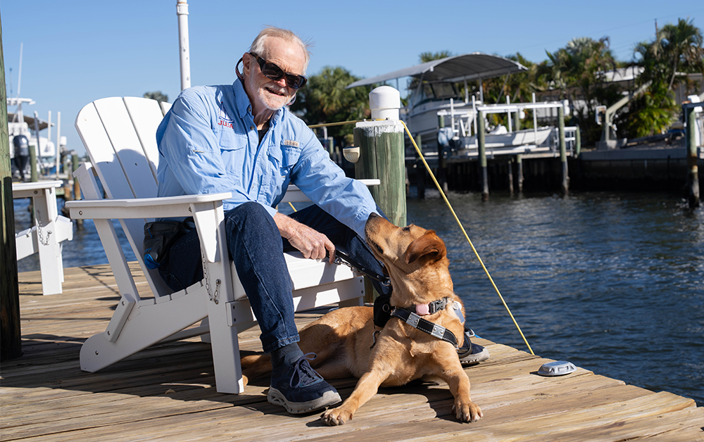 A man sits in a white chair on a boat dock while his yellow Labrador guide dog lays in front of him.