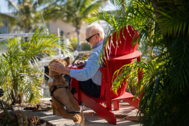 A man sits in a red porch chair and pets his yellow Labrador guide dog that looks up at him.