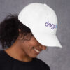 The right side profile of a white dad hat with the Dogs Inc logo embroidered in purple and blue.