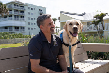 John Alvarez sits with his yellow Labrador service dog on an outdoor bench.