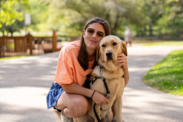 A female crouches and hugs her yellow Labrador guide dog.