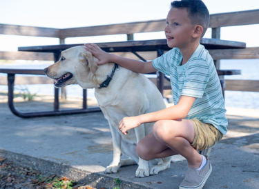 A young boy squats next to his Skilled Companion Dog outside in front of a picnic table.