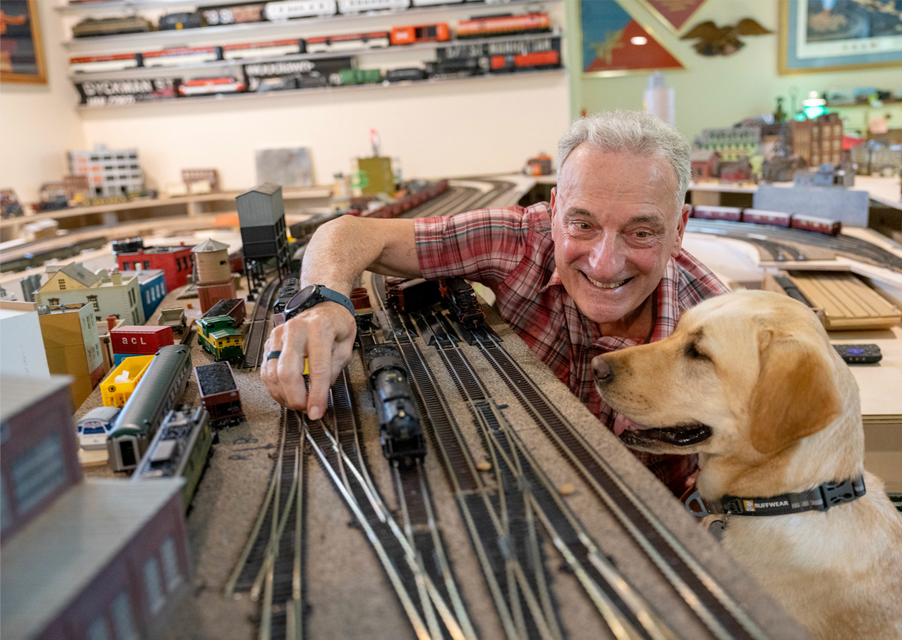 A man adjusts a model train on its track as his yellow lab guide dog watches on