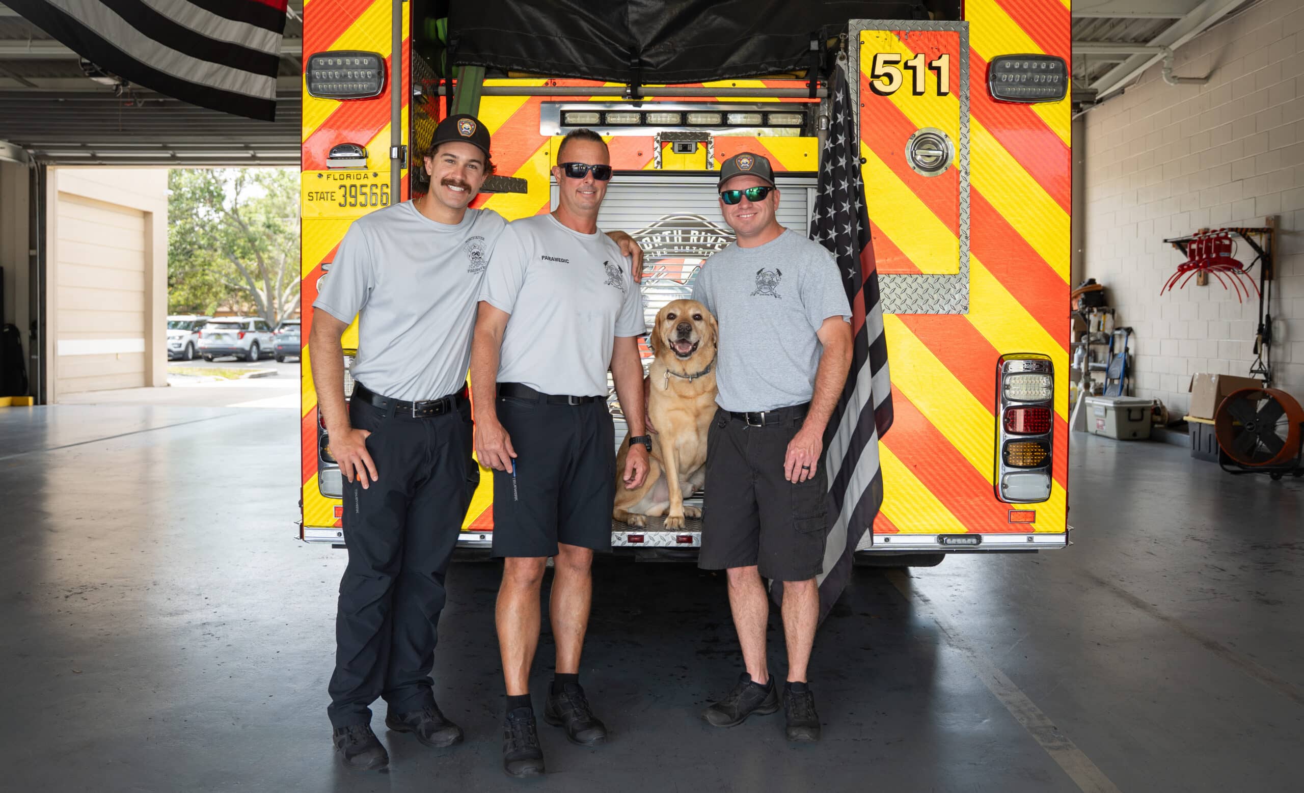 A trio of male firefighters smile and stand at the back of a fire truck with a yellow Labrador.