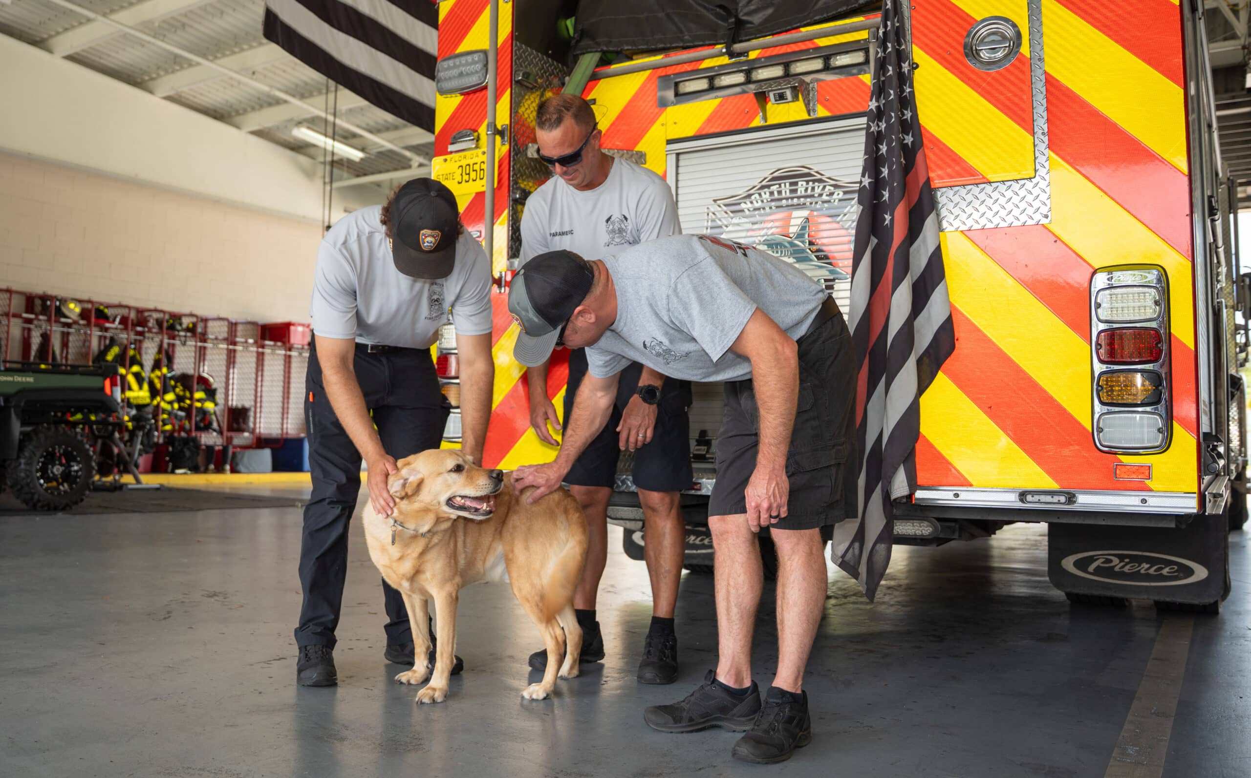 A trio of male firefighters stand and pet a yellow Labrador at the back of a fire truck.