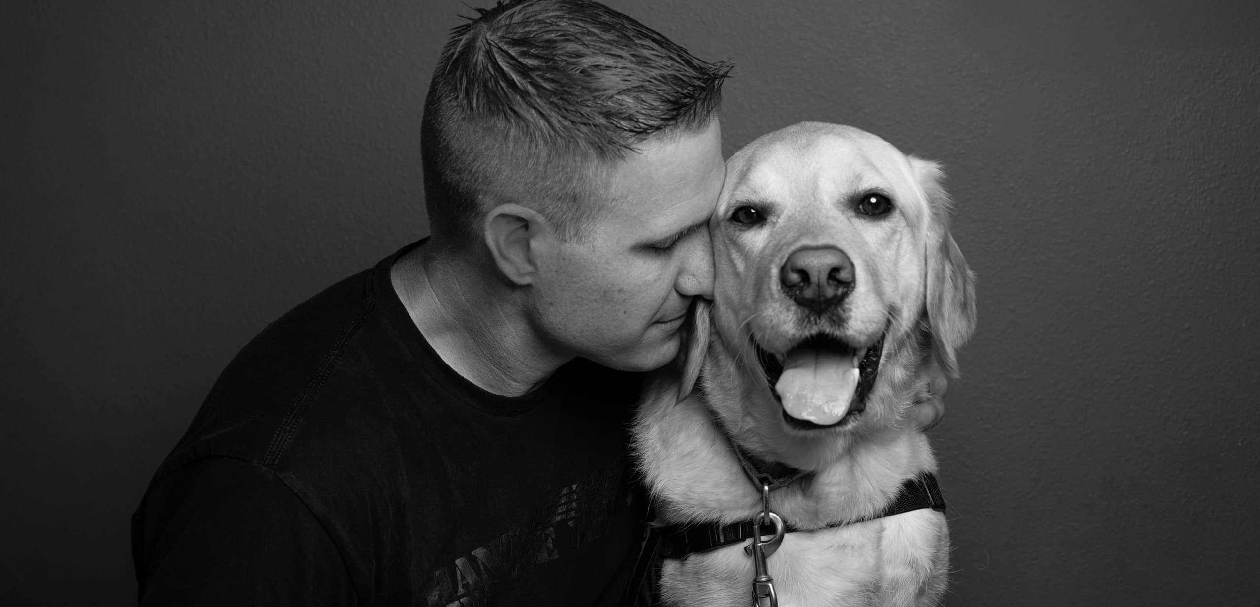 A black and white photo of a man who wears a black t-shirt and rests his head against a yellow Labrador service dog.