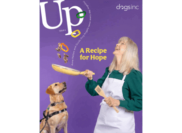 White text reads Up and Dogs Inc. A woman stands in front of a purple background and uses a yellow pan to toss vegetables into the air. A yellow Labrador guide dog sits next to her.