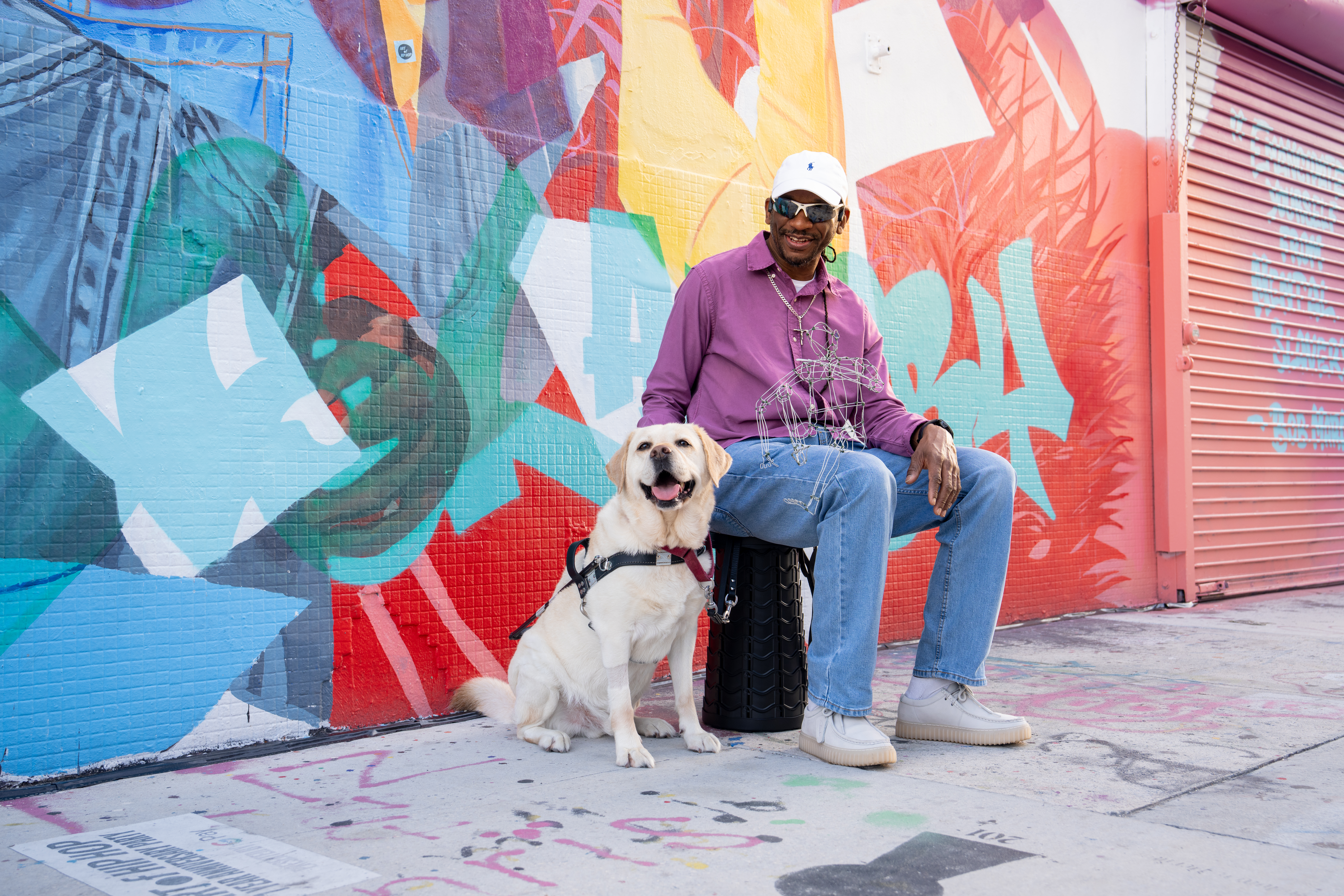 A man sits next to a yellow Labrador guide dog who wears a harness. The man smiles and pets the dog as they sit in front of a colorful, abstract mural.