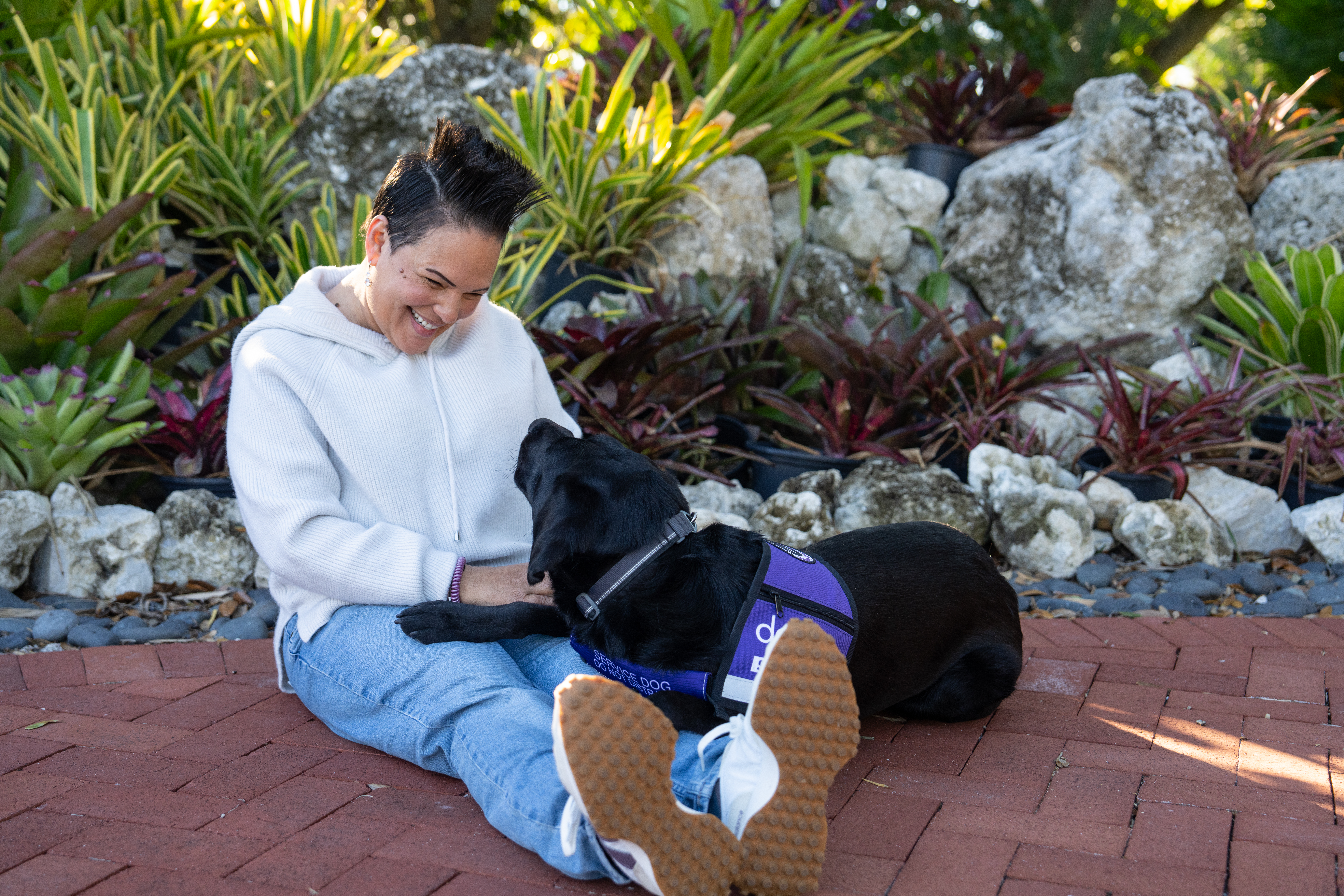 A woman wears a white sweater and sits on a brick paved path next to a black Labrador service dog wears a purple vest with white text that reads, "Dogs Inc Service Dog." The woman smiles at the dog.