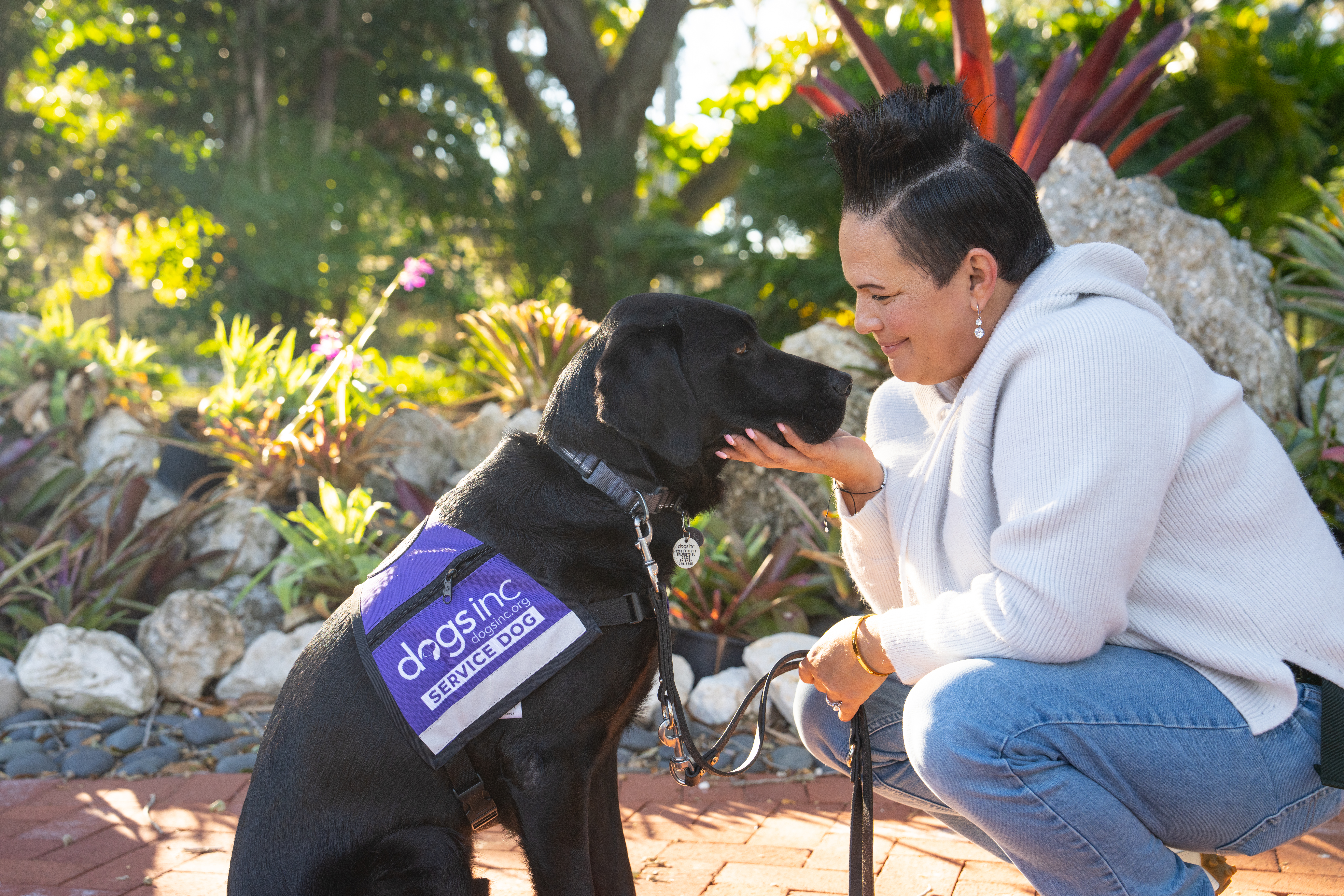A woman wears a white sweater and crouches next to a black Labrador service dog wears a purple vest with white text that reads, "Dogs Inc Service Dog." The woman holds the dog's face and smiles.