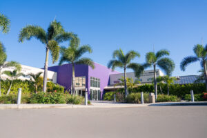 A purple and cream colored building with palm trees surrounding it and a blue sky in the background.
