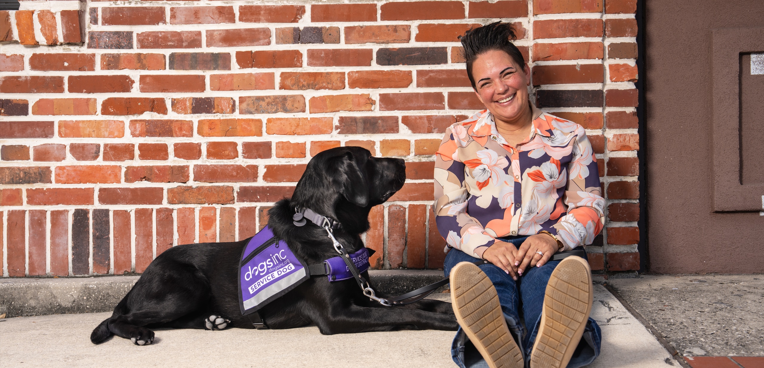 A woman and her black Labrador service dog sit in front of a brick wall. The dog wears a purple vest that says "Dogs Inc Service Dog"