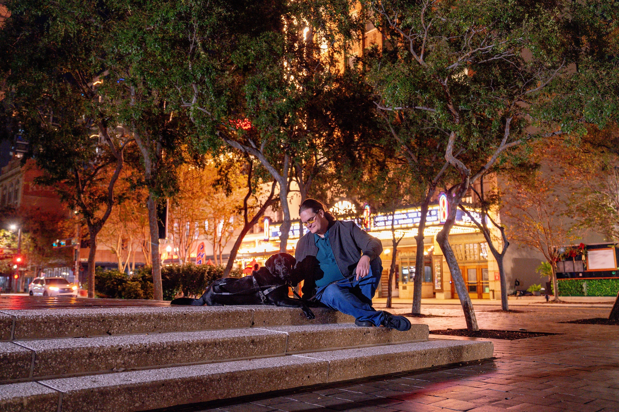 A man sits on steps in front of a theatre next to his black guide dog.