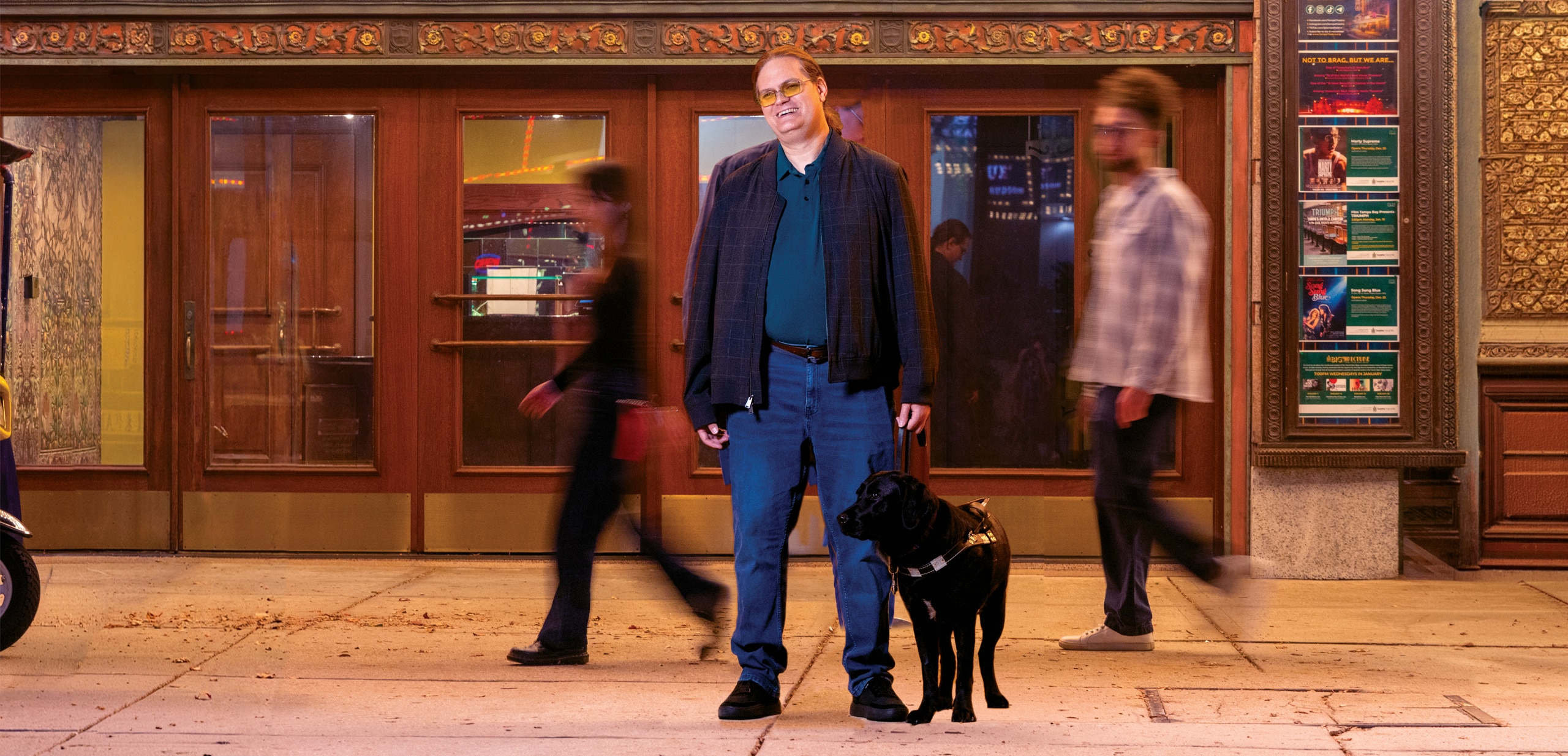 A man and his black Labrador guide dog stand in front of a theatre. The dog wears a black guide dog harness.
