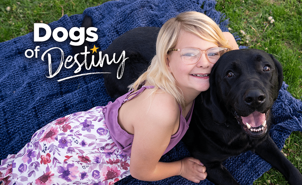 A girl and her black Labrador skilled companion dog lay in the grass on a blue blanket. White text reads, "Dogs of Destiny."