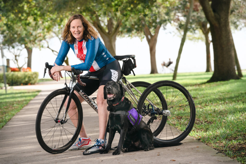 A cyclist on her bike sits next to a Black Lab wearing a purple "Dogs Inc" vest at a sunny park setting.