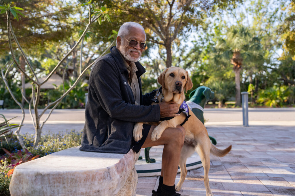 A man sits on a bench outside Dogs Inc campus while a Labrador Retriever in a purple "Dogs Inc Service Dog" vest stands next to him with two paws placed on top of his leg.