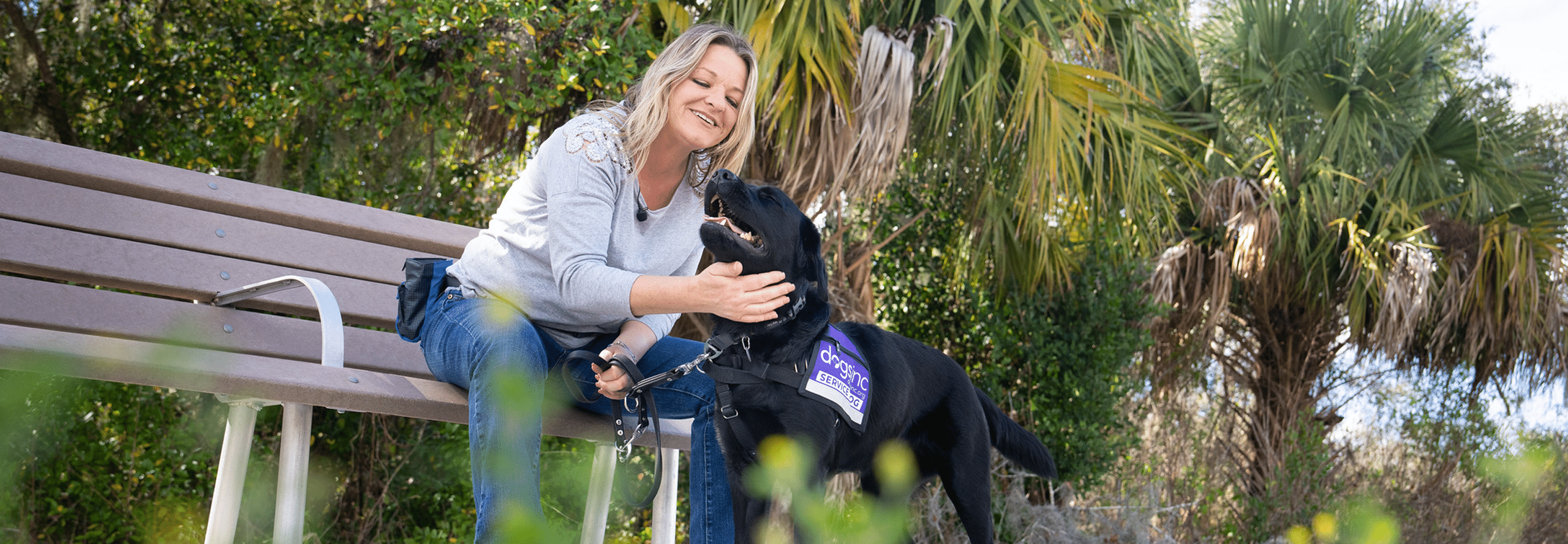 A woman sits on a park bench affectionately petting a Black Lab wearing a purple "Dogs Inc Service Dog" vest while holding the dog’s leash and petting the dog.