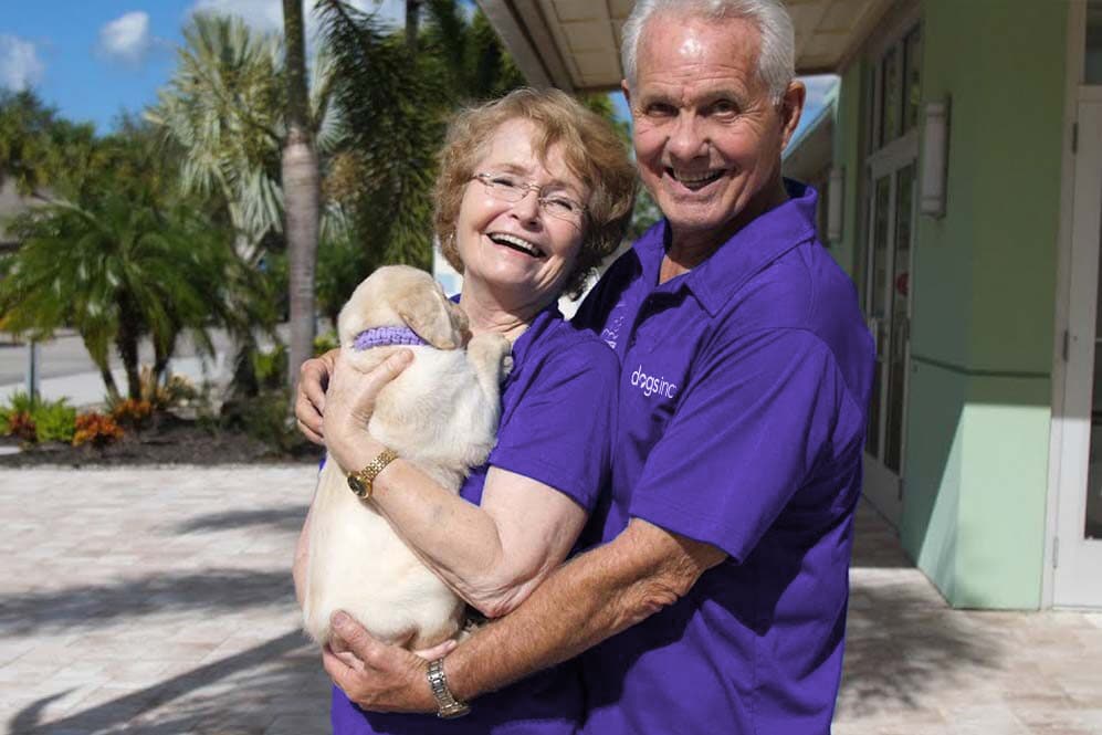 A man and a woman wearing matching purple "Dogs Inc" shirts joyously hold a Labrador Retriever puppy outdoors.