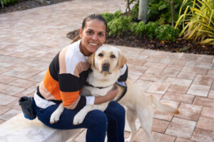 A Labrador Retreiver with a purple "Dogs Inc Service Dog" vest sits beside a smiling woman on an outdoor bench.