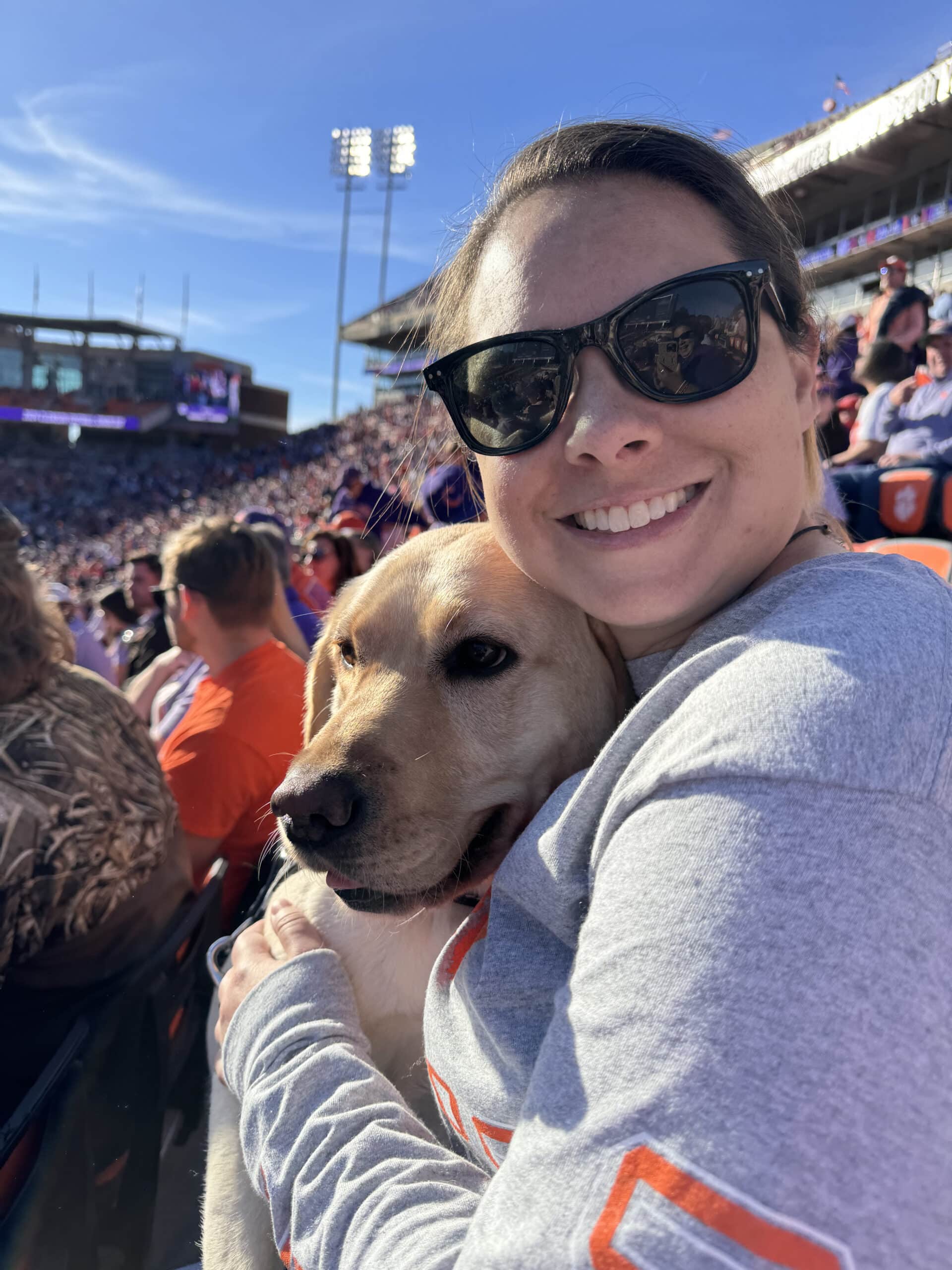 A happy woman is accompanied by a Labrador Retriever at a crowded stadium for an outdoor sporting event.