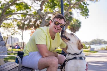 A Yellow Labrador guide dog gleefully sits next to its handler on a park bench on a waterfront path.
