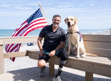 A Yellow Labrador service dog and a man with a prosthetic leg sit on a bench at the beach in front of the American flag.