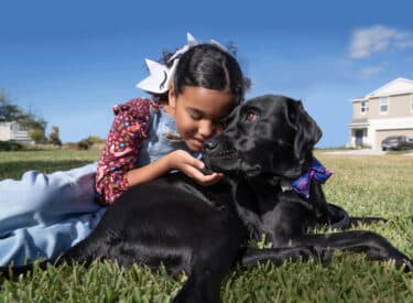 A young girl and a Black Labrador service dog lie on the grass together near a suburban street.