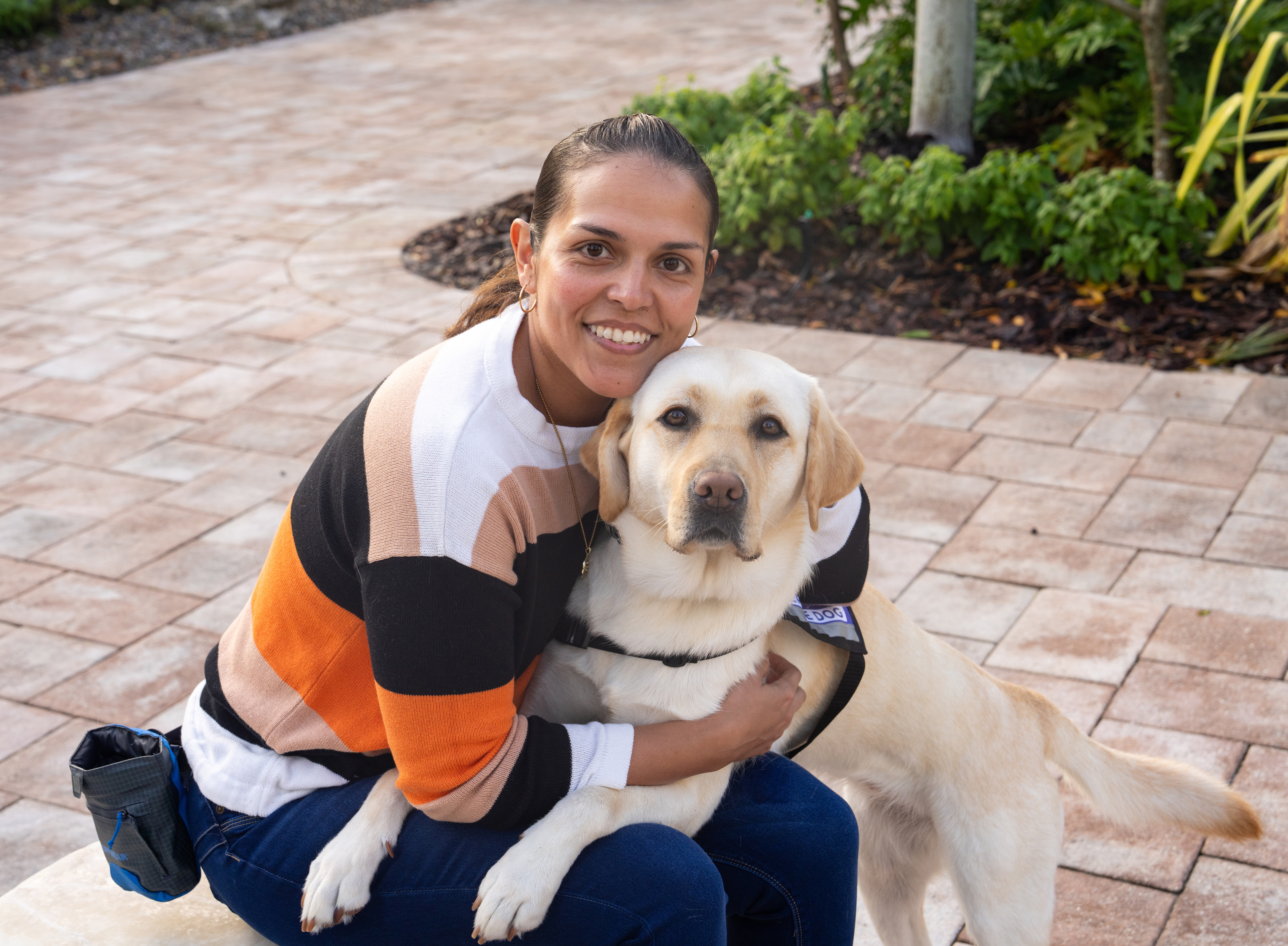 A Yellow Labrador Retriever service dog wearing a purple vest sits beside a smiling woman on an outdoor bench.