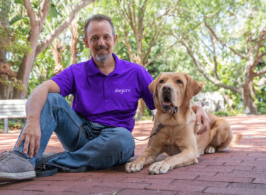 A man wearing a Dogs Inc collared shirt sits with a Labrador Retriever outside on a brick path on a sunny day.