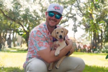 A man holds a yellow lab puppy wearing a purple bandana in his arms outside on the grass in a sunny park setting.