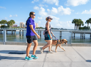 A Dogs Inc pack member member assists a woman walking with a yellow lab guide dog along a waterfront path on a sunny day.