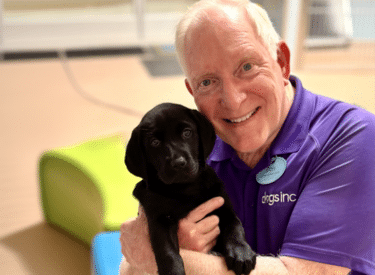 A happy volunteer for Dogs Inc holds a black lab puppy in his arms at an indoor facility.